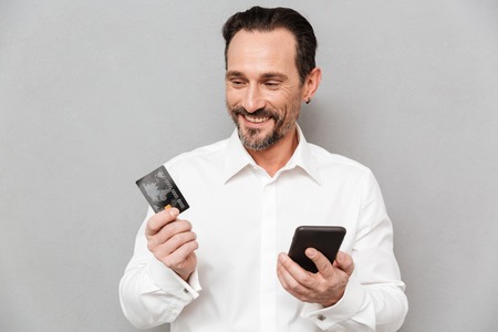 Portrait of a confident mature man dressed in shirt holding mobile phone and a credit card over gray...