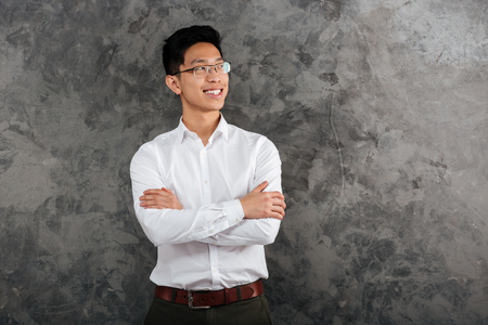 Portrait of a smiling asian man dressed in shirt standing with arms folded and looking away over gray backgroundの写真素材