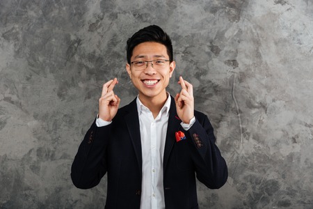 Portrait of a cheerful young asian man dressed in suit holding fingers crossed for good luck over...