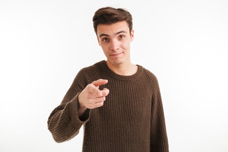 Portrait of a confident young man in sweater pointing finger at camera isolated over white...