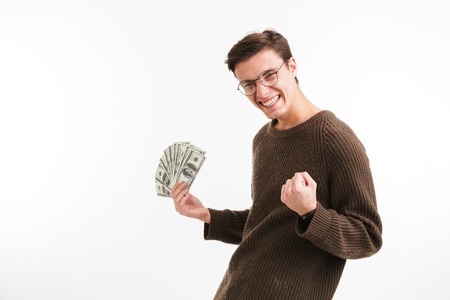 Portrait of a happy young man in sweater holding money banknotes and celebrating isolated over white...