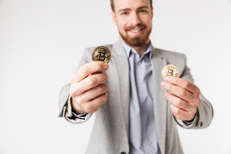 Close up of a smiling young man dressed in shirt and jacket showing two golden bitcoins isolated over white backgroundの写真素材