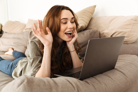 Photo of young happy woman with brown hair lying on sofa in living room and looking on camera of laptop while speaking on videoの写真素材