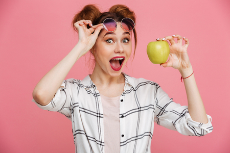 Portrait of a cheerful young girl in sunglasses showing green apple over pink backgroundの写真素材