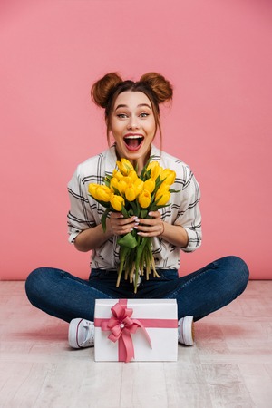 Portrait of a happy young girl holding yellow tulips and present box while sitting on a floor with crossed legs isolated over pink backgroundの写真素材