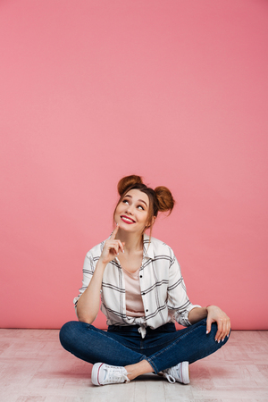 Portrait of a dreamy young girl sitting on a floor with crossed legs and looking up at copy space isolated over pink backgroundの写真素材