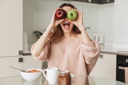 Portrait of a joyful young woman having healthy breakfast while sitting at the table in a kitchen at home in the morningの写真素材