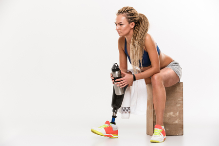 Image of young disabled sports woman with prosthesis sitting isolated over white background. Looking aside drinking water.の写真素材