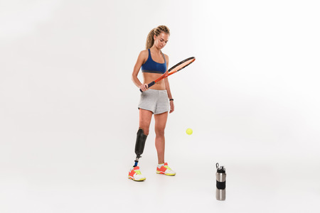 Portrait of a concentrated young disabled sportswoman with leg prosthesis doing exercises with tennis racket isolated over white backgroundの写真素材