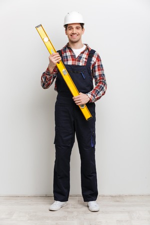 Full length image of Happy male builder in protective helmet holding level tool and looking at the...