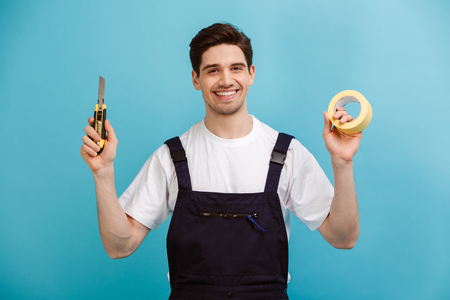 Smiling male builder holding cutter and adhesive tape while looking at the camera over blue backgroundの写真素材