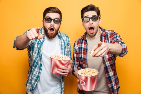 Portrait of a two shocked young men in 3d glasses eating popcorn while standing and pointing fingers...