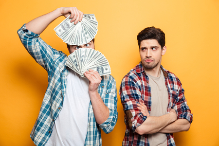 Funny bearded man having fun with money while standing near his displeased friend over yellow backgroundの写真素材