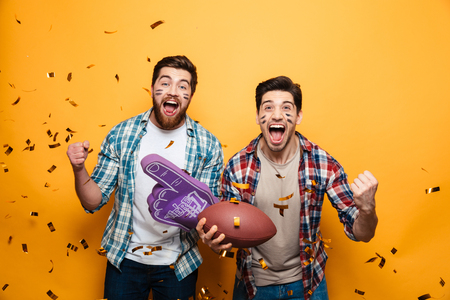 Portrait of a two excited young men holding rugby ball and foam glove while celebrating under confetti rain isolated over yellow backgroundの写真素材