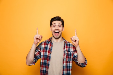 Portrait of a joyful young man pointing fingers up at copy space isolated over yellow background
