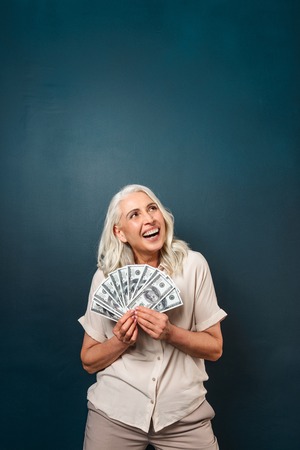 Image of mature happy old woman standing isolated over dark blue background wall looking aside holding money.の写真素材