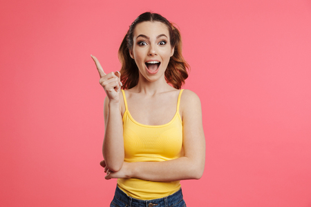 Portrait of an excited young girl dressed in summer clothes pointing finger up and looking at camera isolated over pink backgroundの写真素材