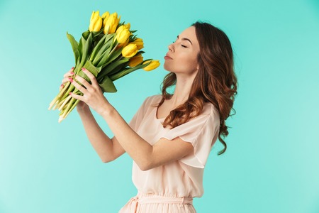 Portrait of a pretty young girl in dress holding yellow tulips bouquet isolated over blue backgroundの写真素材