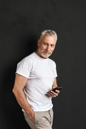 Picture of happy handsome mature old man standing isolated over black wall. Looking aside using...
