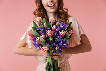 Cropped image of a smiling young girl in dress holding big bouquet of irises and tulips isolated over pink backgroundの写真素材