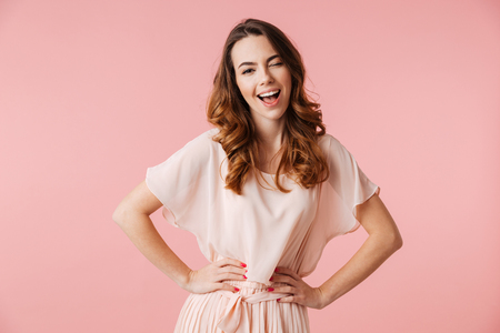 Portrait of a cheerful young girl in dress looking at camera and winking isolated over pink backgroundの写真素材