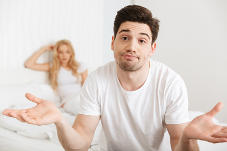Image of young loving couple in bed indoors at home quarrel. Focus on confused man.の写真素材