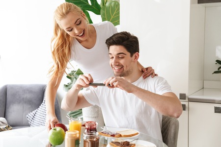 Portrait of a happy young couple in love taking a photo with mobile phone while having tasty breakfast at the table in a kitchenの写真素材