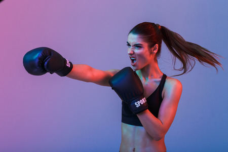 Photo of strong willed woman 20s in sportswear and black boxing gloves throwing punches and fighting with opponent isolated over dark backgroundの写真素材