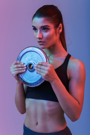 Photo of serious strong young sports woman make exercises with gym barbell disk isolated over purple wall background. Looking aside.の写真素材