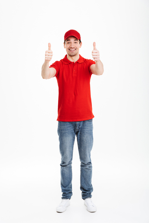 Image of a cheerful young delivery man in red cap standing with thumbs up isolated over white background.の写真素材