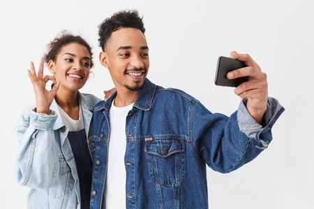 Happy african couple in denim shirts having fun together while making selfie on smartphone over grey backgroundの写真素材