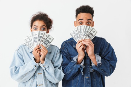 Surprised african couple in denim shirts covering their faces with money while looking at the camera over grey backgroundの写真素材
