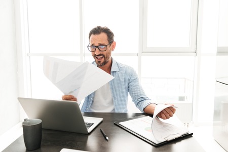 Frustrated mature man working with documents while sitting with laptop computer at the table at home