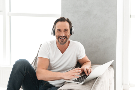 Joyful mature man listening to music with headphones while sitting on a sofa at home with laptop computerの写真素材