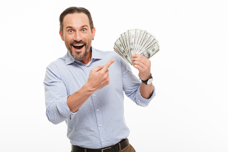 Portrait of an excited mature man dressed in shirt pointing finger at bunch of money banknotes isolated over white backgroundの写真素材