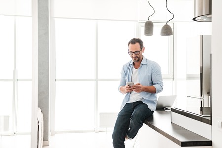 Smiling mature man using mobile phone while leaning on a kitchen table at homeの写真素材