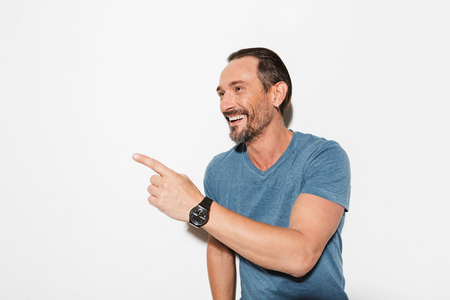 Portrait of a happy mature man dressed in t-shirt pointing finger away at copy space isolated over white backgroundの写真素材