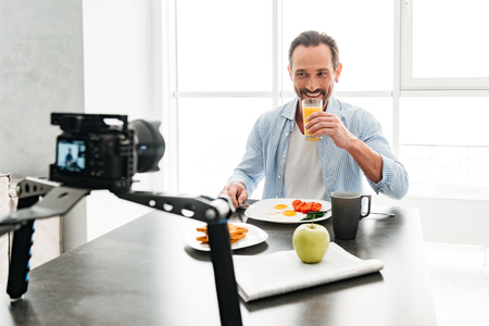 Handsome mature man recording video food blog about cooking on dslr camera in kitchen at homeの写真素材