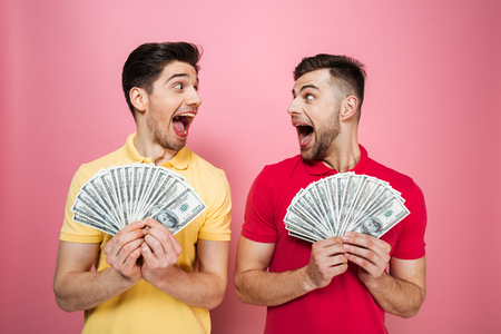 Portrait of an excited gay male couple holding money banknotes and celebrating isolated over pink backgroundの写真素材