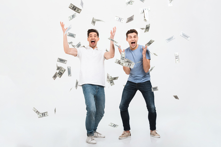 Full length portrait of two happy young men standing under money banknotes shower and celebrating success isolated over white backgroundの写真素材