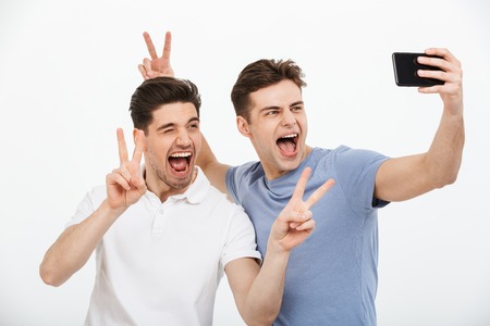 Portrait of two happy young men showing peace gesture while taking a selfie isolated over white backgroundの写真素材