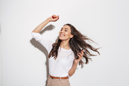 Portrait of a happy asian businesswoman holding mobile phone and dancing isolated over white backgroundの写真素材