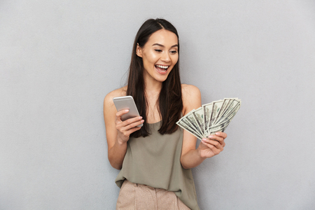 Portrait of a cheerful asian woman holding money banknotes and using mobile phone isolated over gray backgroundの写真素材