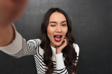 Happy brunette woman in sweater making selfie while winks with eye and looking at the camera over black backgroundの写真素材