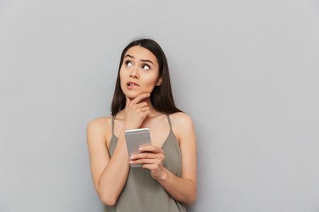 Portrait of a pensive asian woman holding mobile phone and looking away isolated over gray backgroundの写真素材