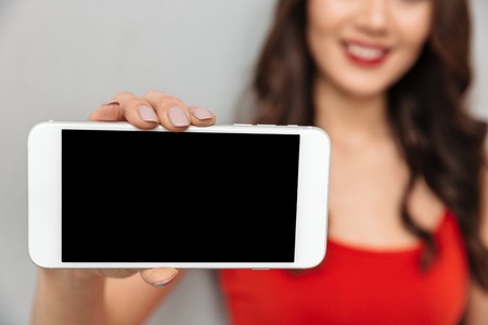 Cropped image of Smiling brunette woman in casual clothes showing blank smartphone screen at camera over grey background. Focus on phoneの写真素材