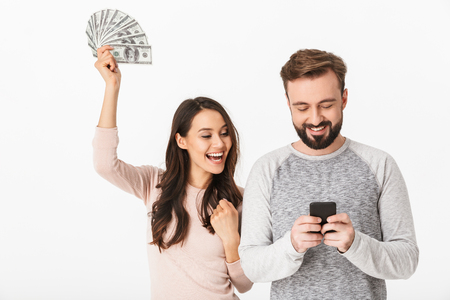 Photo of happy young loving couple standing isolated over white background holding money using mobile phone.の写真素材