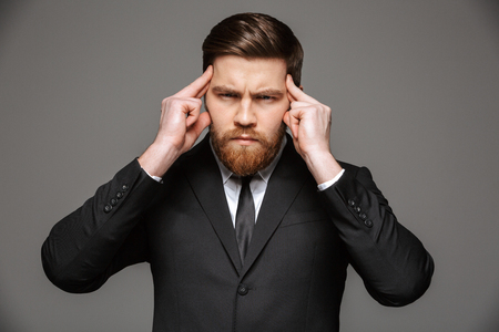 Portrait of a concentrated young businessman dressed in suit with fingers on his temple isolated over gray backgroundの写真素材