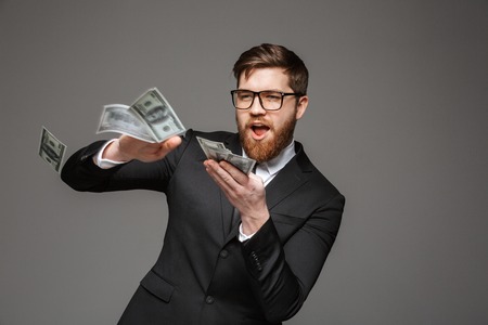 Portrait of a happy young businessman throwing out money banknotes isolated over gray background