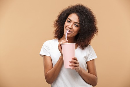 Portrait of a cheerful young african woman drinking from a plastic cup isolated over beige backgroundの写真素材